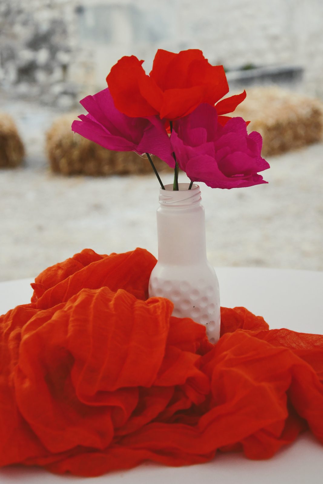 Fleur en papier fuchsia dans un vase blanc, posée sur un voile orange